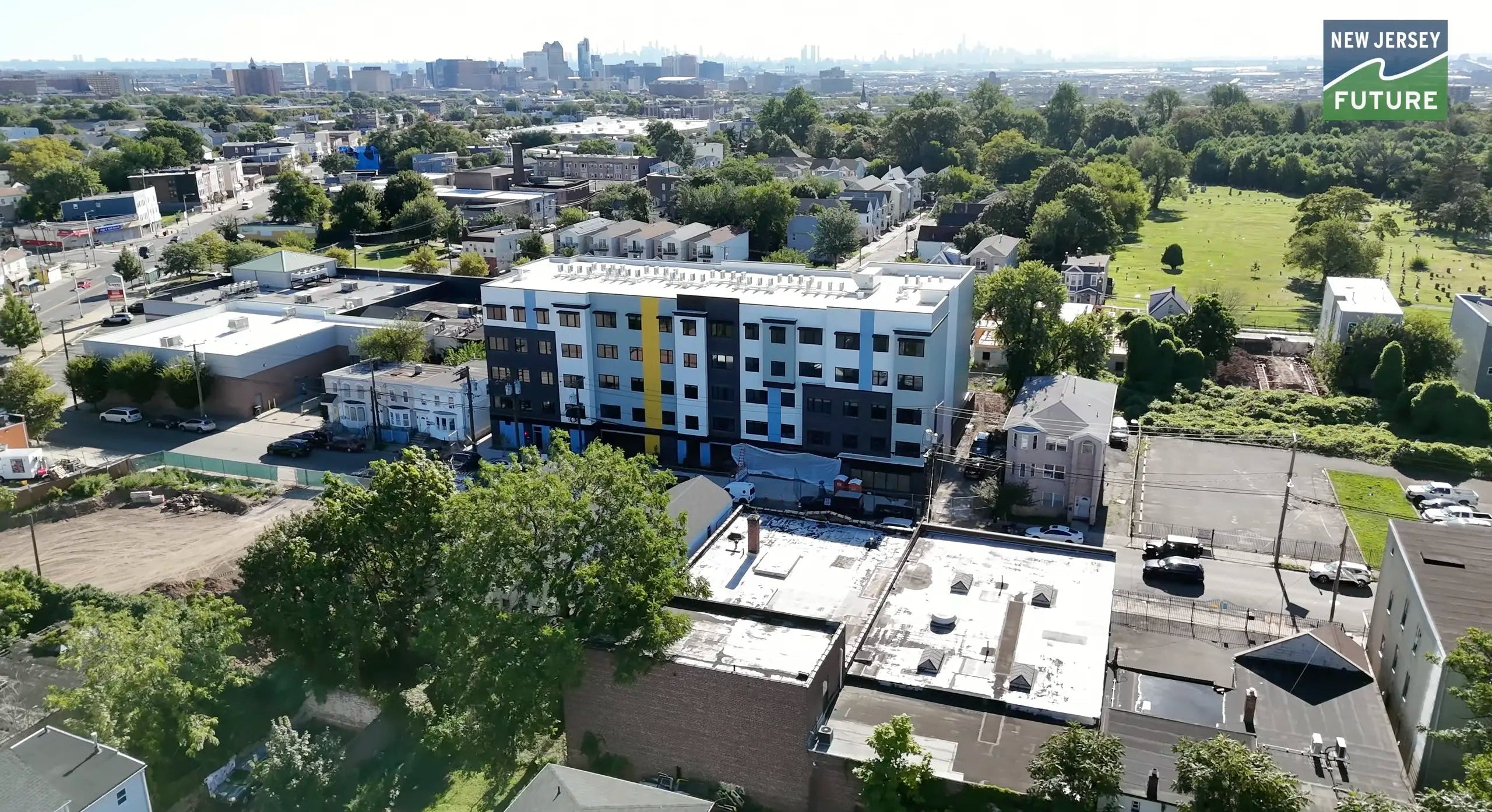 Aerial view of an urban area with a building under construction, trees, and a city skyline in the distance.